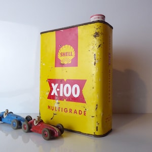May include: A vintage yellow Shell X-100 Multigrade oil can with red detailing and the Shell logo. Two toy racing cars, one blue and one red, are positioned in front of the can. The can has a red and white cap.