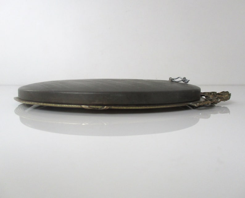 May include: Oval-shaped picture frame with a dark gray backing and a gold-colored metal frame. The frame has a decorative design on the bottom edge and a metal hook on the back for hanging.