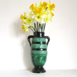 Pode incluir: Um vaso de cerâmica verde e preto com duas alças, cheio de narcisos amarelos e brancos.