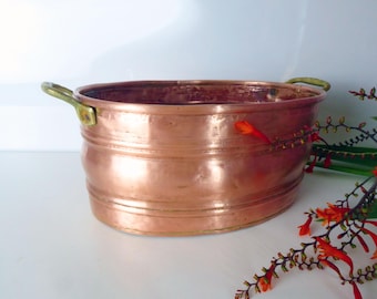 Può includere: Un contenitore ovale in metallo color rame con due manici in ottone. Il contenitore presenta fasce orizzontali sul corpo. Lo sfondo è bianco, con rametti di fiori arancioni. Per scopi decorativi.
