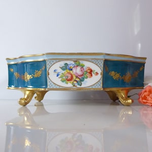 May include: A teal and white porcelain planter with gold accents and floral decorations. The planter has three gold feet and a scalloped edge.