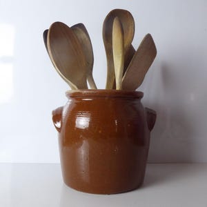 May include: A brown glazed ceramic crock with two side handles containing a collection of various wooden spoons and spatulas. The kitchen utensil holder has a smooth, glossy finish and a cylindrical shape, holding several light-colored wooden tools.