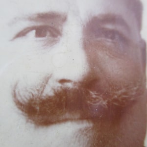 May include: A sepia-toned photograph of a man's face with a mustache. The man is looking directly at the camera. The photo is faded and has a vintage look.