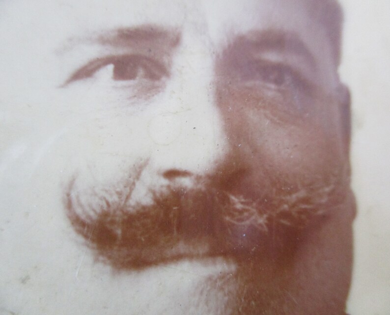 May include: A sepia-toned photograph of a man's face with a mustache. The man is looking directly at the camera. The photo is faded and has a vintage look.