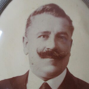 May include: A sepia-toned portrait of a man with a mustache, wearing a suit and tie. The photo is framed in a gold oval frame.
