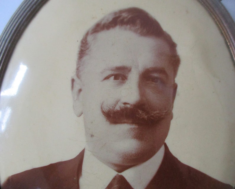May include: A sepia-toned portrait of a man with a mustache, wearing a suit and tie. The photo is framed in a gold oval frame.