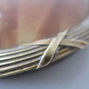 May include: Close-up of a round, brown and white agate cameo with a gold metal frame. The frame is made of multiple thin strips of metal that are woven together in a criss-cross pattern.