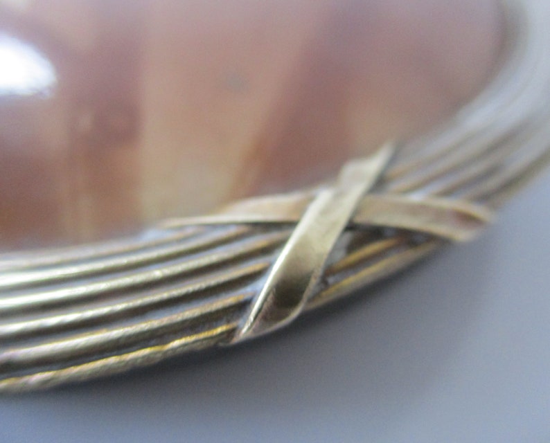 May include: Close-up of a round, brown and white agate cameo with a gold metal frame. The frame is made of multiple thin strips of metal that are woven together in a criss-cross pattern.