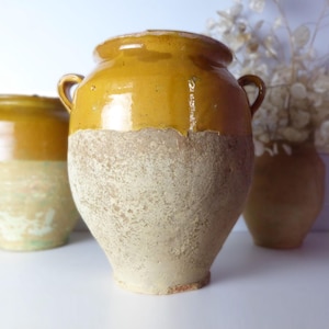 May include: Three antique ceramic jars with a rustic, aged appearance. The jars are two-toned, with a mustard yellow top and a textured, off-white lower half. One jar has handles. The background includes a vase with dried flowers.