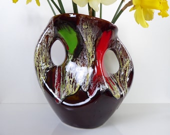 May include: A brown ceramic vase with abstract red, green, and white glaze. The vase has two cut-out handles and a unique, asymmetrical shape.