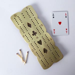 May include: A brass cribbage board with decorative cutouts of card suits and a playing card showing the Ace of Hearts. Three matches are next to the board. The board is a light gold color.