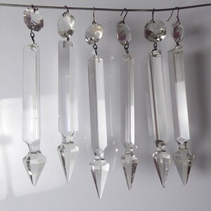 以下が含まれることがあります： ワイヤーに吊るされた6つの透明なクリスタルシャンデリアプリズム。各プリズムは、ファセット加工された上部と、先端に向かって細くなる長い長方形のボディを備えています。クリスタルは白い背景に吊るされています。