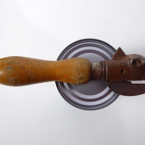 May include: A vintage can opener with a wooden handle and a metal head shaped like an animal. The head has a small hole in the side and a small, round eye.
