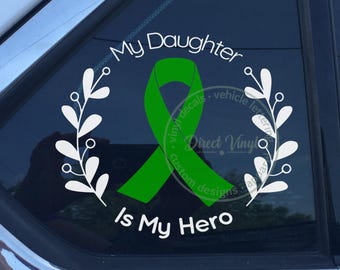 Può includere: Un adesivo in vinile bianco per auto con un nastro di sensibilizzazione verde centrale circondato da una corona di foglie bianche. Il testo My Daughter è arcuato sopra il nastro e il testo Is My Hero è posizionato sotto in bianco.