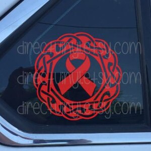 Può includere: Adesivo in vinile rosso raffigurante un nastro all'interno di un disegno a nodo celtico, applicato al finestrino di un'auto. L'adesivo riporta il testo "dinecviny.com" ripetuto verticalmente. L'auto è bianca con finiture cromate.