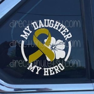 Puede incluir: Una calcomanía de vinilo blanco con el texto "My Daughter My Hero" en un círculo. Una cinta amarilla con una flor blanca está en el centro del círculo.