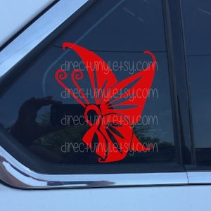 Può includere: Adesivo a forma di farfalla rossa su un finestrino di un'auto. La farfalla ha un design stilizzato con accenti neri.