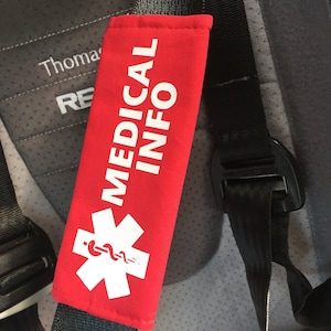 May include: A bright red seat belt cover featuring the white text MEDICAL INFO and a white Star of Life symbol with a central serpent and staff icon. The rectangular fabric sleeve is wrapped around a black nylon seat belt strap.
