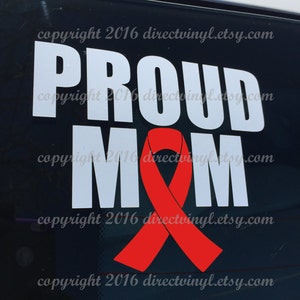 Può includere:  Adesivo in vinile bianco con le parole "Proud Mom" e un nastro rosso. L'adesivo è su uno sfondo nero.