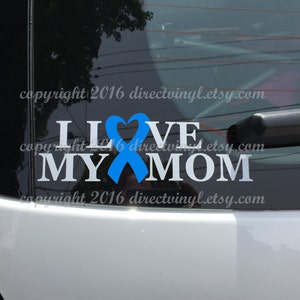 May include: White vinyl decal with the words "I Love My Mom" and a blue ribbon with a heart in the center. The decal is on the back window of a car.