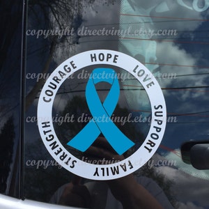 May include: A white circular car decal with a light blue ribbon in the center. The words "COURAGE", "HOPE", "LOVE", "SUPPORT", "FAMILY", and "STRENGTH" are printed around the ribbon. The decal is on a car window.