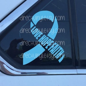 May include: A light blue ribbon with the words "Love Hope Faith" printed on it. The ribbon is on a car window.