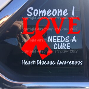 Può includere: Nastro rosso per la consapevolezza delle malattie cardiache con il testo "Someone I LOVE NEEDS A CURE" e le parole "Heart Disease Awareness" sotto.