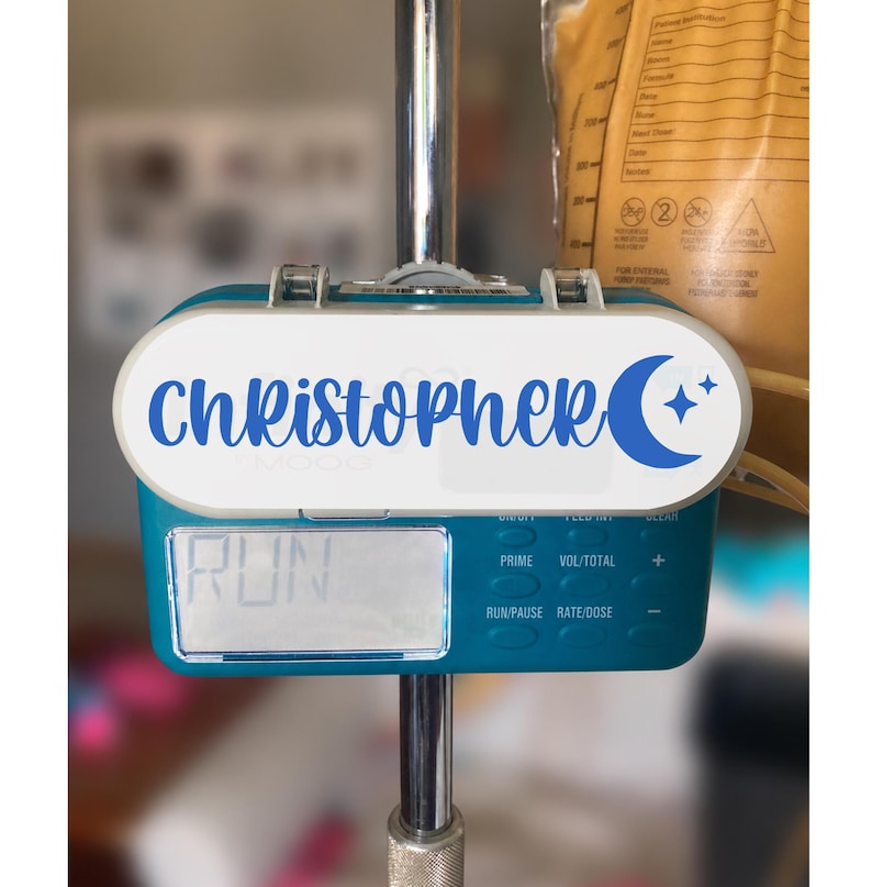 K&ouml;nnte beinhalten: Eine t&uuml;rkisfarbene medizinische Ern&auml;hrungspumpe an einer Metallstange mit einem wei&szlig;en ovalen Etikett, das den blauen Schriftzug "Christopher" sowie eine Mondsichel und zwei kleine Sterne zeigt. Das Display zeigt das Wort "RUN" an.