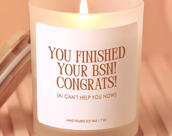 May include: A lit, frosted glass candle with a wooden lid. The candle has a white label with the text "YOU FINISHED YOUR BSN! CONGRATS! (AI CAN'T HELP YOU NOW)". The candle is hand-poured soy wax, 7 oz.