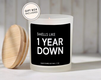 May include: A white frosted glass candle with a black label that reads "SMELLS LIKE 1 YEAR DOWN." The candle has a wooden lid and is labeled "HAND POURED SOY WAX | 7 OZ." A gift box is included.
