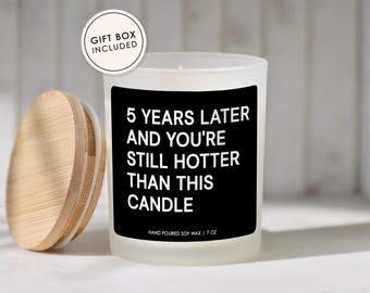 May include: A frosted glass candle with a black label featuring the white text 5 YEARS LATER AND YOU'RE STILL HOTTER THAN THIS CANDLE. The candle includes a light wood lid and a circular tag stating GIFT BOX INCLUDED. It contains 7 oz of soy wax.