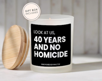 May include: A frosted glass candle with a black label featuring the white text LOOK AT US, 40 YEARS AND NO HOMICIDE and HAND POURED SOY WAX | 7 OZ. A round bamboo lid sits to the left, and a circular badge states GIFT BOX INCLUDED.