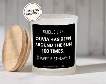 May include: A frosted glass candle with a black label that reads: SMELLS LIKE OLIVIA HAS BEEN AROUND THE SUN 100 TIMES. (HAPPY BIRTHDAY!) HAND POURED SOY WAX | 7 OZ. The candle has a light-colored wooden lid and a small circular tag stating GIFT BOX INCLUDED.