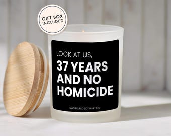 May include: A frosted glass candle with a black label featuring the white text: LOOK AT US, 37 YEARS AND NO HOMICIDE. The candle includes a light wood lid and a circular tag stating GIFT BOX INCLUDED. The label also reads HAND POURED SOY WAX | 7 OZ.
