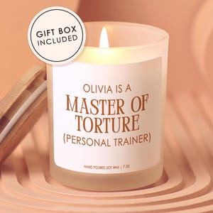 May include: A frosted glass candle with a bamboo lid, featuring a white label with brown text that reads: OLIVIA IS A MASTER OF TORTURE (PERSONAL TRAINER) HAND POURED SOY WAX | 7 OZ. A circular badge on the top left states GIFT BOX INCLUDED.