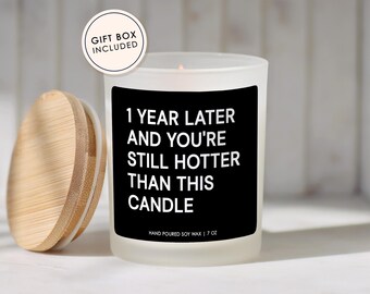May include: A white frosted glass candle with a black label that reads "1 YEAR LATER AND YOU'RE STILL HOTTER THAN THIS CANDLE." The candle has a wooden lid and is labeled "HAND POURED SOY WAX | 7 OZ." A gift box is included.