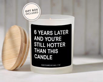 May include: A frosted glass candle with a black label featuring the white text 6 YEARS LATER AND YOU'RE STILL HOTTER THAN THIS CANDLE and HAND POURED SOY WAX | 7 OZ. A round bamboo lid sits to the left, and a circular badge reads GIFT BOX INCLUDED.