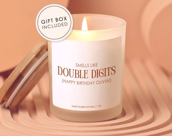 May include: A lit, frosted glass candle with a wooden lid, labeled "Smells Like Double Digits (Happy Birthday Olivia!)". The candle is 7 oz and comes in a gift box. The candle sits on a peach-colored, layered surface.