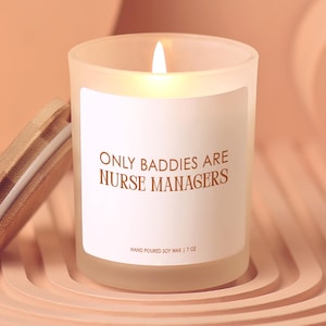 May include: A lit, frosted glass candle with the text "ONLY BADDIES ARE NURSE MANAGERS" on a white label. The candle has a wooden lid and is labeled "HAND POURED SOY WAX | 7 OZ". The background is a light peach color.