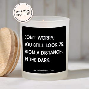 May include: A white frosted glass candle with a black label that reads "DON'T WORRY, YOU STILL LOOK 79. FROM A DISTANCE. IN THE DARK." The candle has a wooden lid and is labeled "HAND POURED SOY WAX | 7 OZ."