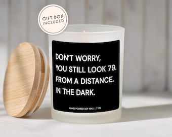 May include: A white frosted glass candle with a black label that reads "DON'T WORRY, YOU STILL LOOK 79. FROM A DISTANCE. IN THE DARK." The candle has a wooden lid and is labeled "HAND POURED SOY WAX | 7 OZ."
