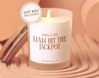 May include: A lit, frosted glass candle with the text "Smells Like Liam Hit The Jackpot" on a white label. The candle has a wooden lid and is labeled as hand-poured soy wax, 7 oz. A circular "Gift Box Included" sticker is in the upper left.