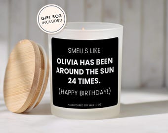 May include: A frosted glass candle with a black label featuring white text that reads: SMELLS LIKE OLIVIA HAS BEEN AROUND THE SUN 24 TIMES. (HAPPY BIRTHDAY!) HAND POURED SOY WAX | 7 OZ. The candle has a light wood lid and a circular tag stating GIFT BOX INCLUDED.