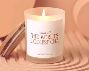 May include: A lit, frosted glass candle with a wooden lid. The candle has a white label with the text "Smells Like The World's Coolest CNA." The candle is hand-poured soy wax and is 7 oz.