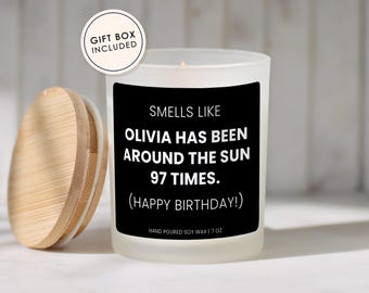 May include: A frosted glass candle with a black label reading SMELLS LIKE OLIVIA HAS BEEN AROUND THE SUN 97 TIMES. (HAPPY BIRTHDAY!) HAND POURED SOY WAX | 7 OZ. A round bamboo lid sits to the left. A circular badge reads GIFT BOX INCLUDED.