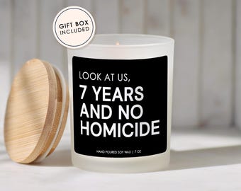 May include: A frosted glass candle with a black label featuring the white text LOOK AT US, 7 YEARS AND NO HOMICIDE and HAND POURED SOY WAX | 7 OZ. A round wooden lid sits to the left, and a circular badge states GIFT BOX INCLUDED at the top left.