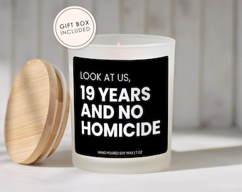 May include: A frosted glass candle with a black label featuring the white text LOOK AT US, 19 YEARS AND NO HOMICIDE, HAND POURED SOY WAX | 7 OZ, and a circular badge stating GIFT BOX INCLUDED. A round wooden lid sits to the left of the candle.