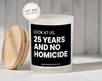 May include: A frosted glass candle with a black label featuring the white text LOOK AT US, 25 YEARS AND NO HOMICIDE and HAND POURED SOY WAX | 7 OZ. A round bamboo lid sits to the left, and a circular badge reads GIFT BOX INCLUDED.