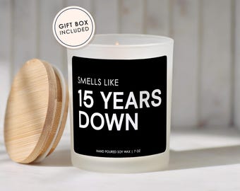 May include: A white candle with a black label that reads "Smells Like 15 Years Down." The candle is in a frosted glass jar with a wooden lid. The text "Hand Poured Soy Wax | 7 oz" is at the bottom.