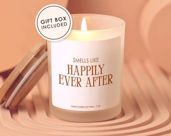 May include: A lit, frosted glass candle with a wooden lid, labeled "Smells Like Happily Ever After." The candle is hand-poured soy wax and is 7 oz. A gift box is included. The background is a soft peach color.